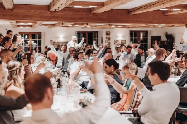 Festliche Hochzeitsgesellschaft hebt gemeinsam die Weingläser zum Toast in einem rustikalen Saal mit Holzbalkendecke. 