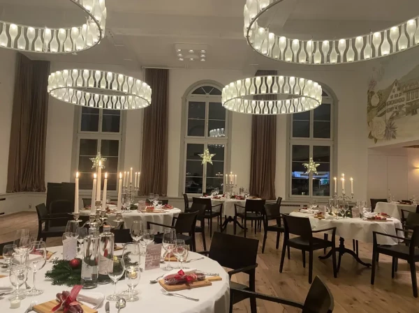 Löwen-Saal bei Nacht mit eindrucksvollen Ringlampen und Weihnachtsdekoration. Runde Tische festlich gedeckt, Kerzenleuchter und leuchtende Sterne an den Bogenfenstern.