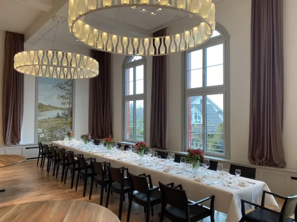 Lange Festtafel mit weissem Tischtuch und roten Blumengestecken. Landschaftsgemälde in historischem Stil und hohe Bogenfenster mit Ausblick.