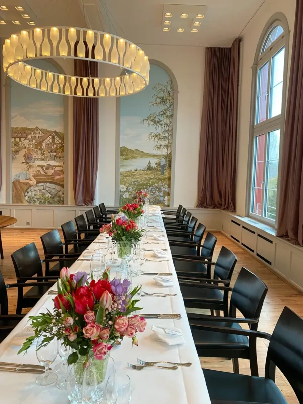 Gedecker Tisch mit farbenfrohen Tulpen- und Rosengestecken vor Wandgemälden in historischem Stil. Ringförmige Deckenleuchte und hohe Bogenfenster prägen den Raum.