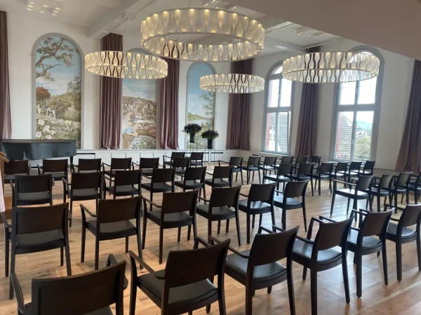 Löwen-Saal mit Reihenbestuhlung für Veranstaltung. Wandgemälde im historischen Stil und moderne Ringlampen prägen den eleganten Raum.