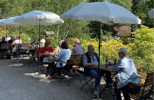 Gemütliche Gartenwirtschaft mit Holzbänken und Sonnenschirmen. Gäste geniessen Erfrischungen inmitten blühender Moorlandschaft.