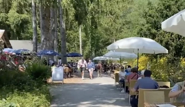 Lebendige Gartenwirtschaft an sonnigem Tag. Besucher flanieren auf dem Kiesweg zwischen Sitzgruppen und üppiger Parkbepflanzung