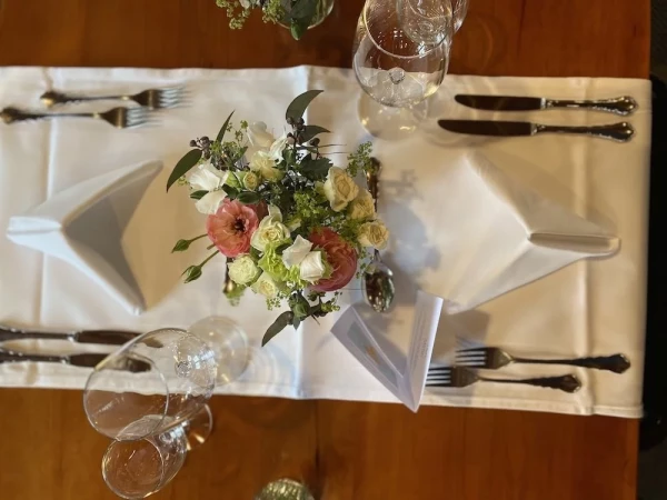 Festlich gedeckter Tisch von oben fotografiert, mit schneeweisser Tischdecke und elegantem Gedeck. Als Mittelpunkt ein zartes Blumenarrangement mit rosa Ranunkeln, cremefarbenen Rosen und frischem Grün in einer Glasvase. 