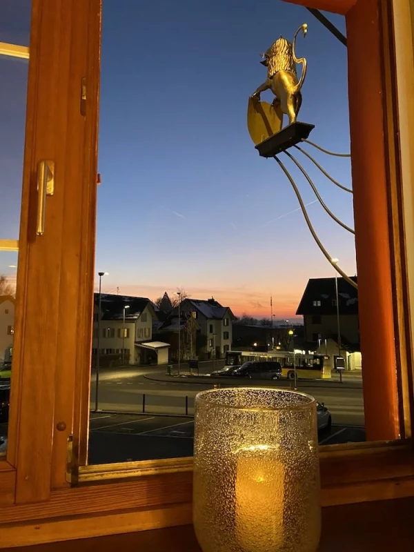 Blick aus dem Restaurant-Löwen-Fenster auf den Postplatz und die Bushaltestelle in der Dämmerung, mit goldenem Löwenemblem und Kerzenlicht im Vordergrund und warmem Abendhimmel über den Dorfhäusern.